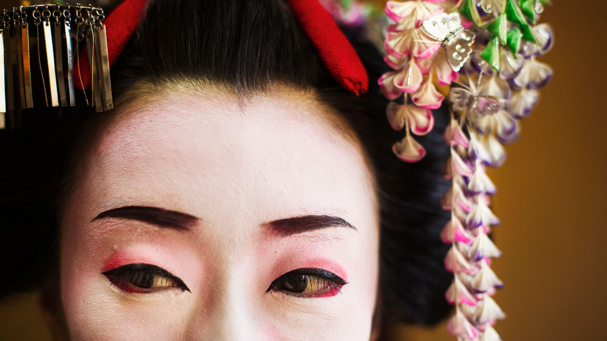 صورة لغييشا يابانية بمكياج أبيض، مع محدِّد عيون أسود وأحمر، وترتدي غطاء رأس مزداناً بالزهور والزخارف.