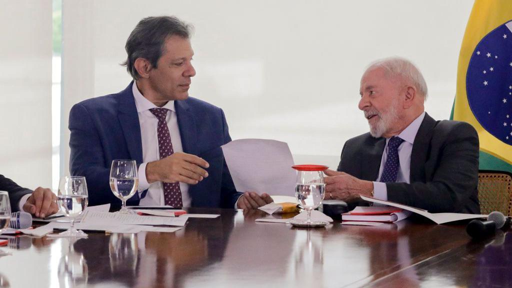Fernando Haddad e Lula conversando e trocando pap&eacute;is em uma mesa de trabalho, com a bandeira do Brasil ao fundo, no canto direito