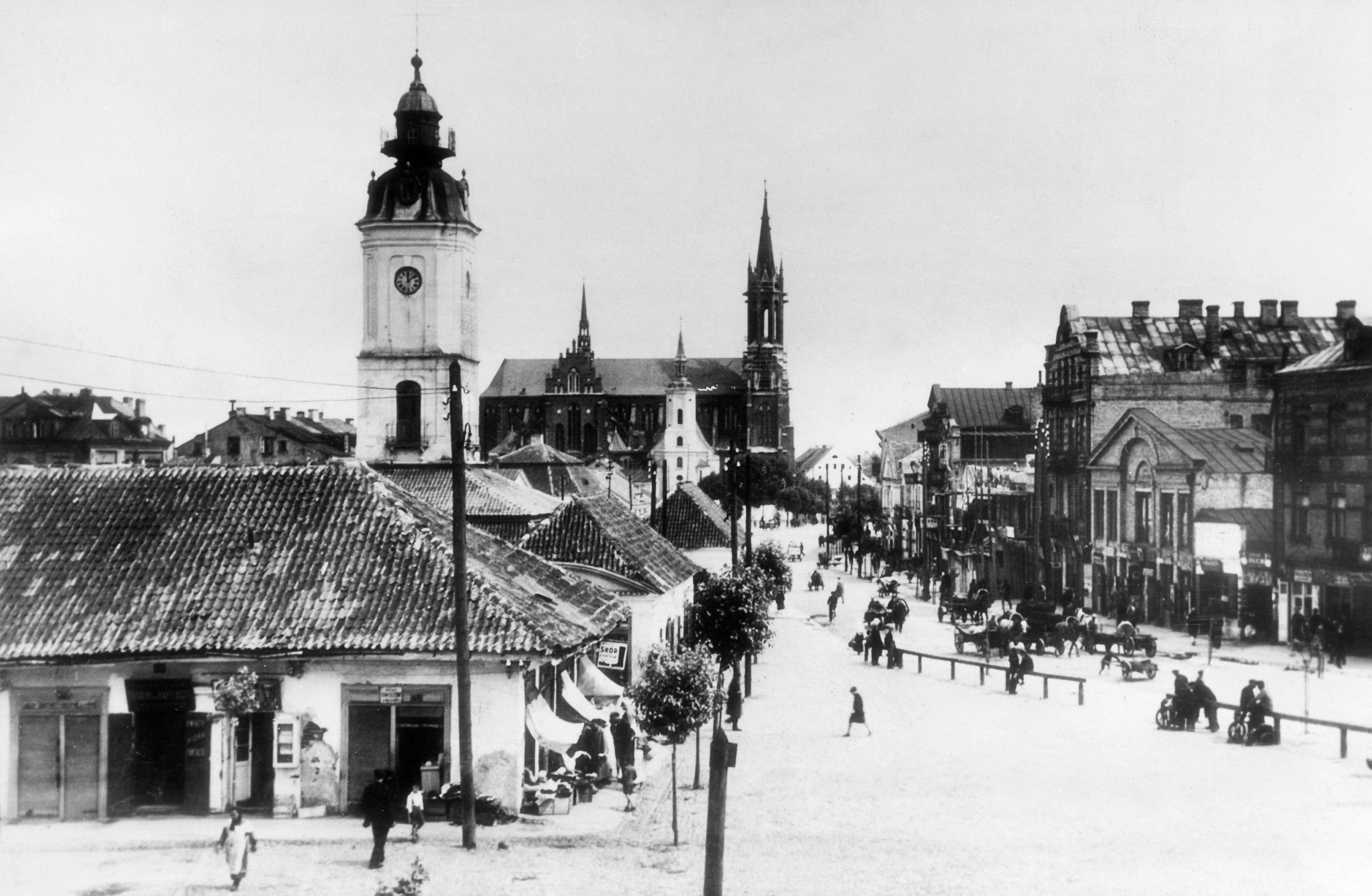 Esquina de uma rua com um pr&eacute;dio de telhado inclinado e uma torre. V&aacute;rias pessoas caminham pela rua, que parece estar coberta por neve. 