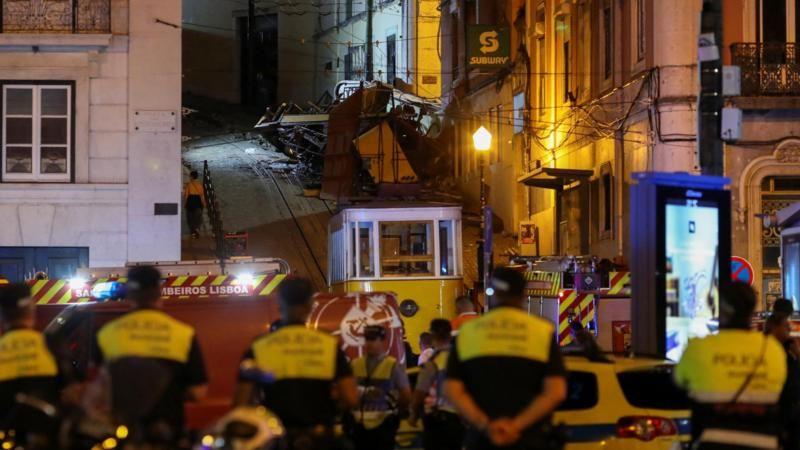 Agentes de policía y equipos de rescate observan el funicular accidentado en una calle empinada de Lisboa durante la noche.