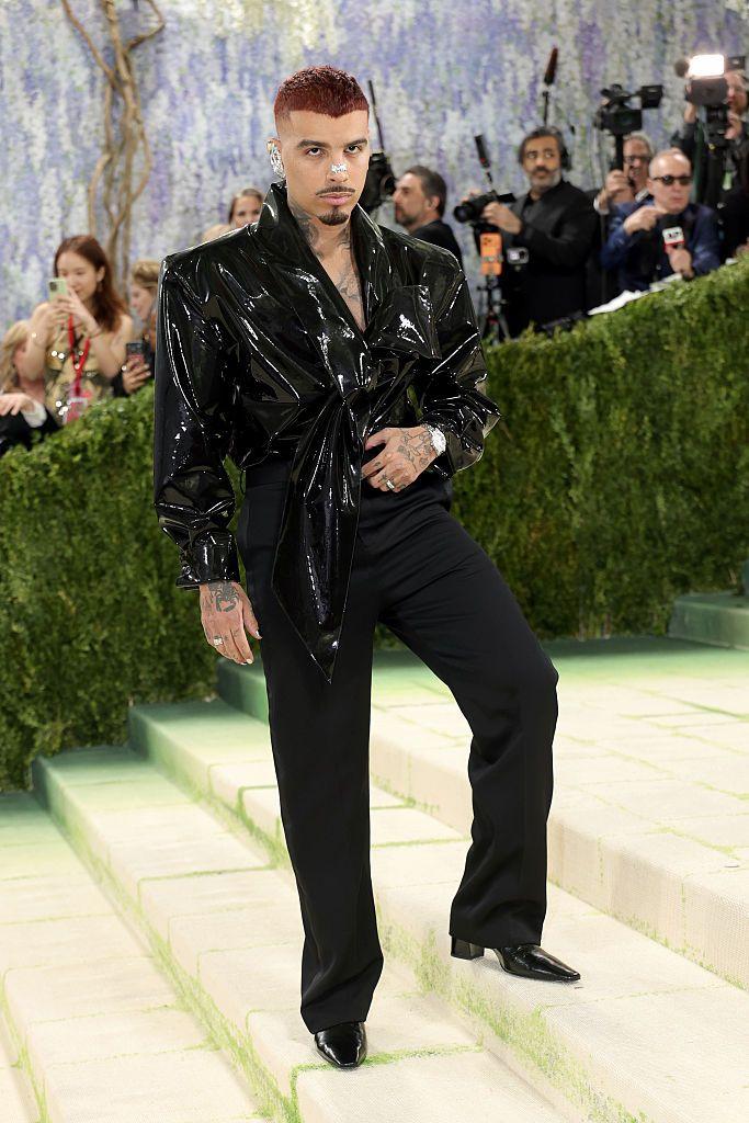 Rauw Alejandro posa en una escalinata verde durante un evento de gala, con un atuendo negro brillante y pantalones haciendo juego.
