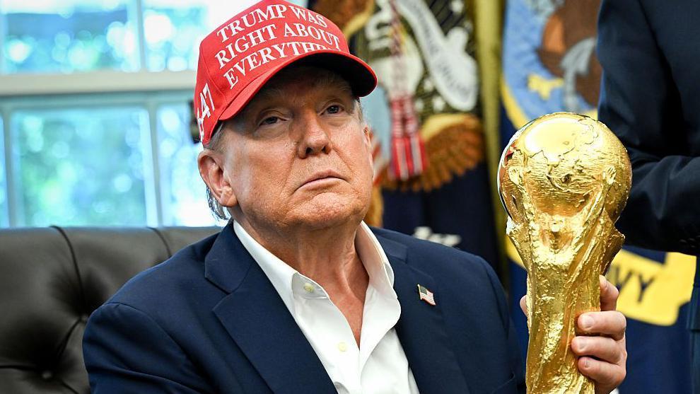 يظهر الرئيس الأمريكي دونالد ترامب، من صدره فما فوق، ممسكاً بكأس العالم لكرة القدم، جالساً على كرسي أسود في المكتب البيضاوي بالبيت الأبيض. يرتدي سترة زرقاء تحمل شارة علم أمريكي على طية صدرها، وقميصاً أبيض مفتوح الياقة، وقبعة بيسبول حمراء كُتب عليها "كان ترامب محقاً في كل شيء". في الخلفية، تظهر نافذة وعلمان، لكنهما غير واضحين.