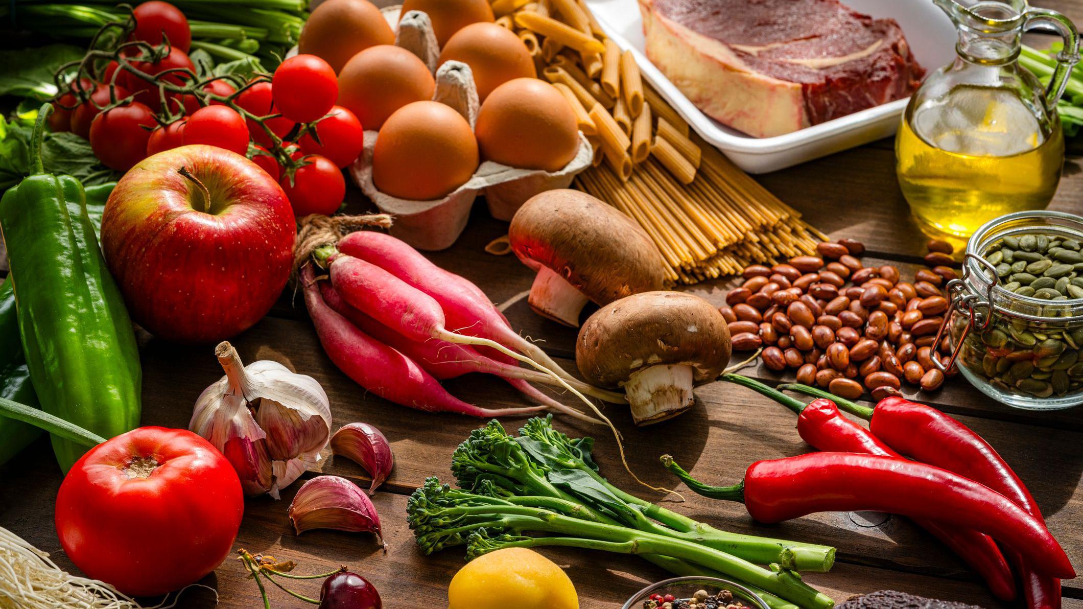 Sobre una mesa de madera se colocan frutas, verduras, semillas, legumbres, carne, pasta, huevos y aceite de cocina.