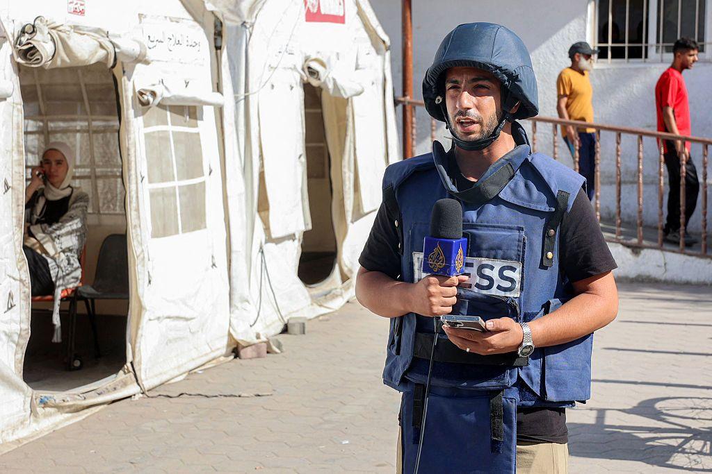 Anas al Sharif con un casco y un chaleco de protección en el que dice "Prensa" hace un reporte a cámara para Al Jazeera, con un micrófono del canal catarí en una mano y un móvil en la otra. Está junto a unas tiendas de campaña y se puede ver a otras personas pasando por detrás de él. 