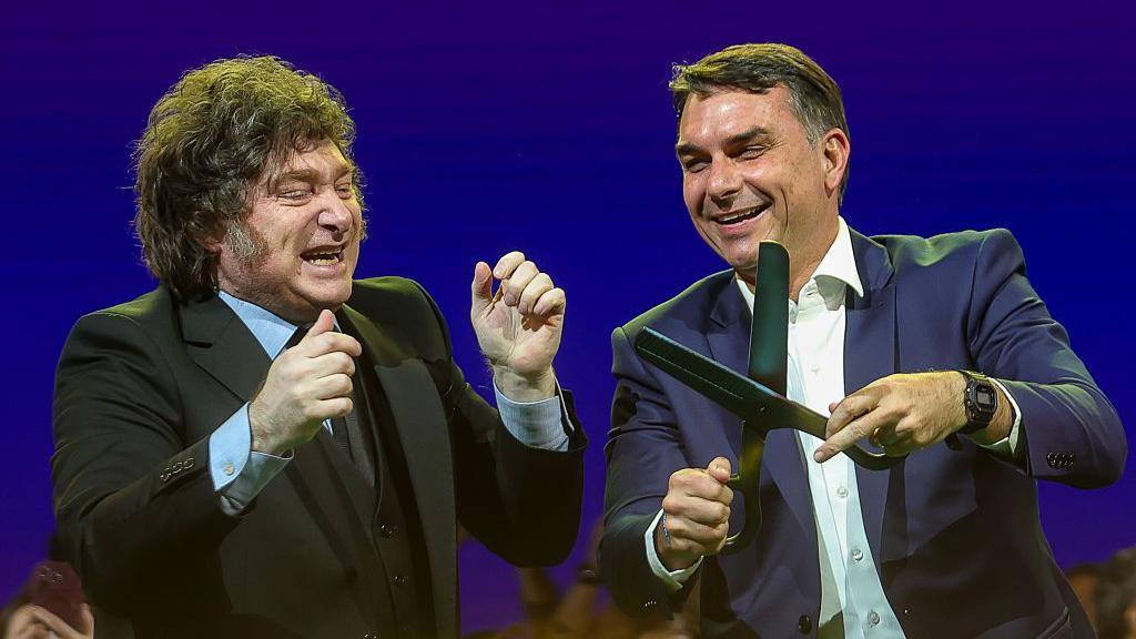 Javier Milei e Fl&aacute;vio Bolsonaro, que segura uma imensa tesoura nas m&atilde;os. 