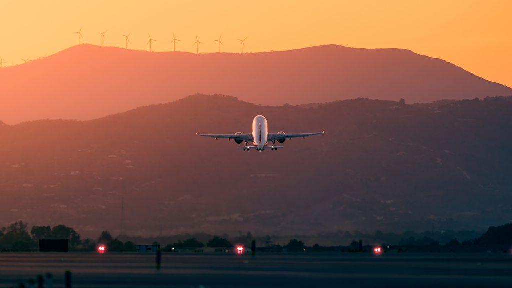 Avião decolando em dia de sol, com montanhas e turbinas eólicas ao fundo