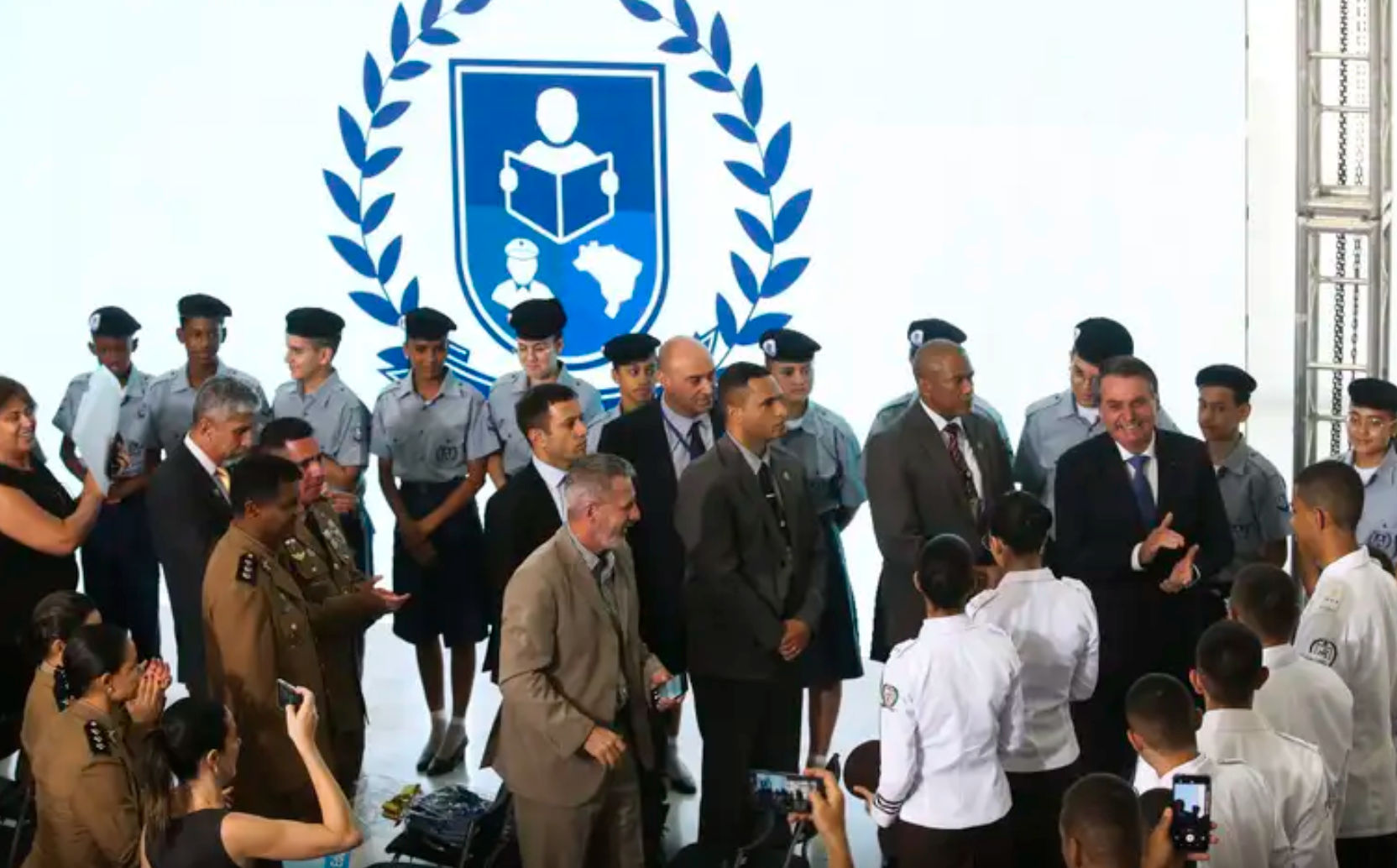 Presidente Jair Bolsonaro rodeado de militares e estudantes, com fundo branco, em evento p&uacute;blico