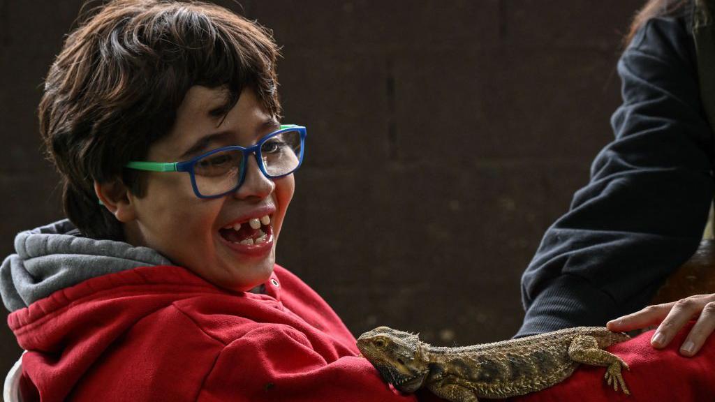 Foto de uma criança de óculos e blusa vermelha com um lagarto em seu braço.