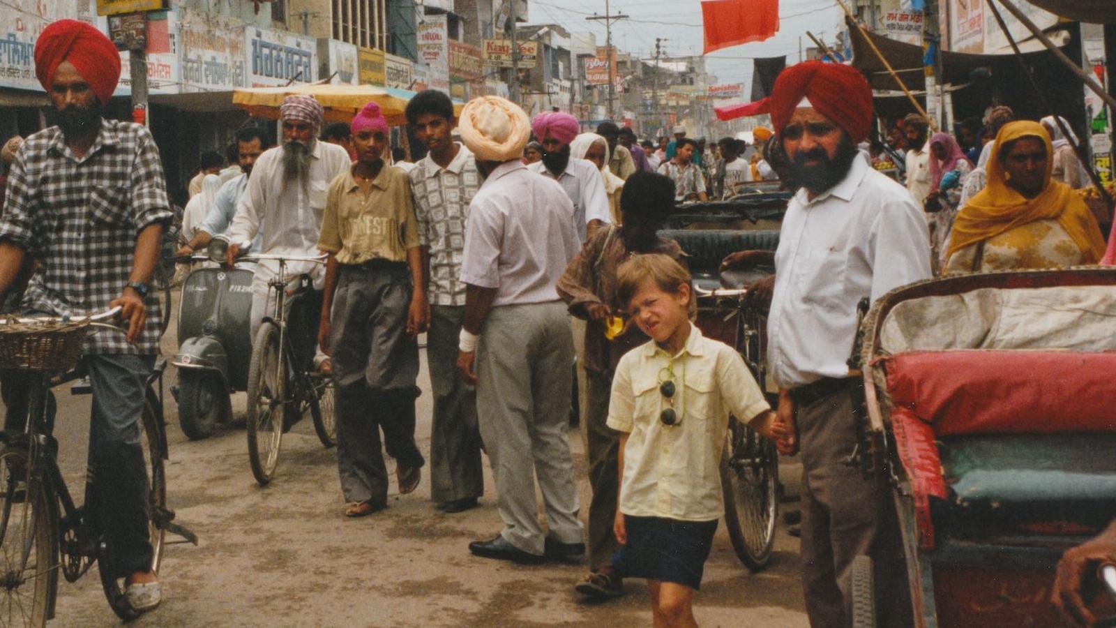 A young boy wearing a white shirt and shorts. He is about six years old and has sunglasses attached to his shirt.He is holding the hand of his Grandad's who is wearing a red turban and has a black beard. He is wearing a light blue shirt and grey trousers with a black belt. They are in India with a lot of people in the background and people riding bikes 