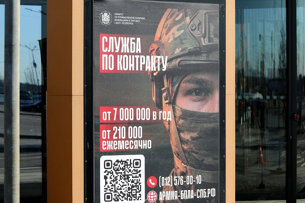 Un cartel de reclutamiento en San Petersburgo muestra la cabeza de un soldado con casco y la boca cubierta. Sobre un fondo rojo, palabras en ruso blancas, números, un número de teléfono y un código QR prometen a los soldados contratados un salario anual de hasta siete millones de rublos, o 210.000 al mes. Una escena callejera se refleja en el cristal negro que rodea el cartel.

