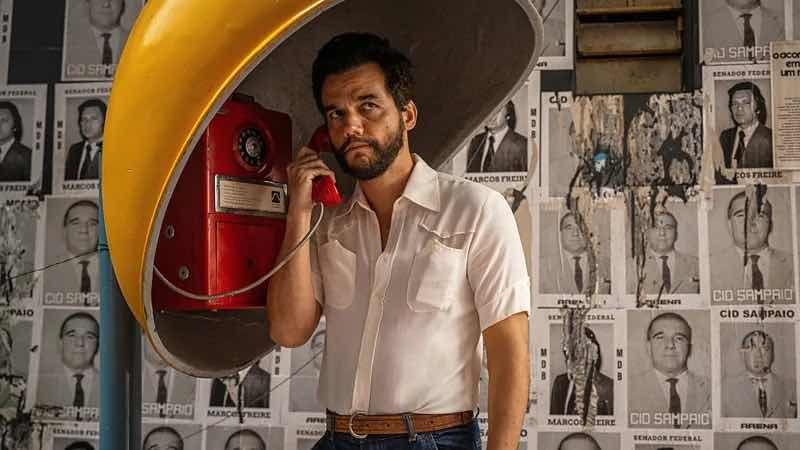 Wagner Moura falando a um telefone na rua. Há vários cartazes atrás