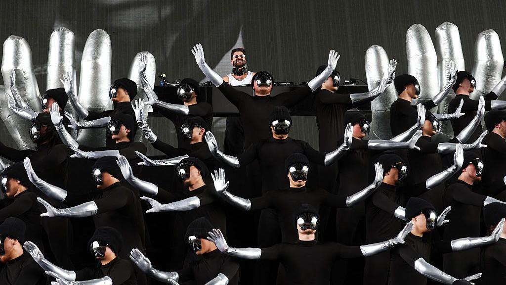 Alok se apresenta ao fundo do palco, cercado por dezenas de performers vestidos de preto, com bra&ccedil;os prateados e m&aacute;scaras espelhadas que ocultam seus rostos. Os dan&ccedil;arinos executam uma coreografia sincronizada com os bra&ccedil;os erguidos em diferentes dire&ccedil;&otilde;es.