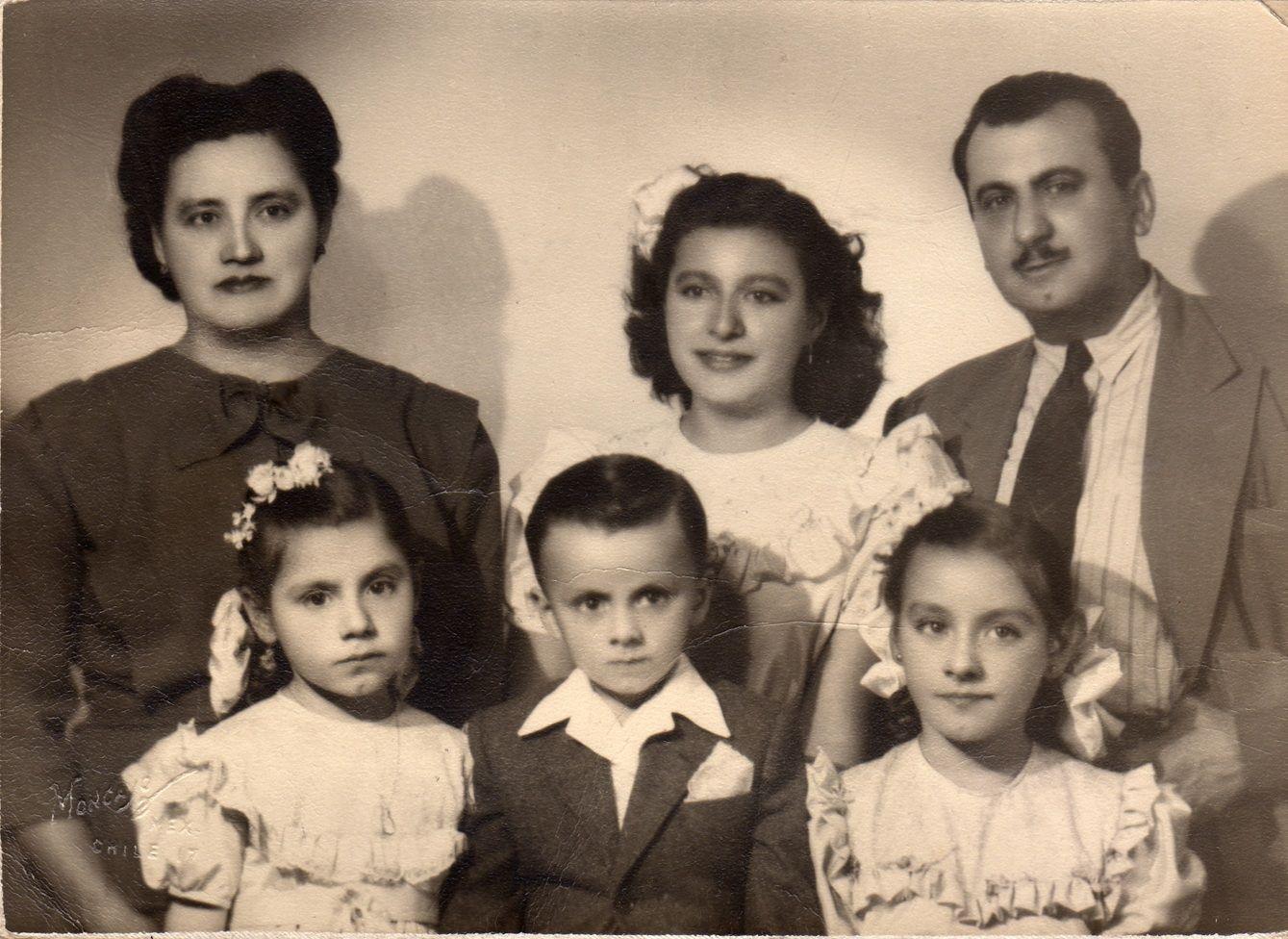 La madre de Alejandra Jáidar, con un vestido oscuro, está parada en la parte izquierda. A su lado y en el centro de la foto está Alejandra, de adolescente, con un vestido blanco y un adorno blanco en la cabeza. A su lado está su padre, vestido de traje y corbata. Delante de ellos están tres niños, uno de ellos es un niño que está en el centro