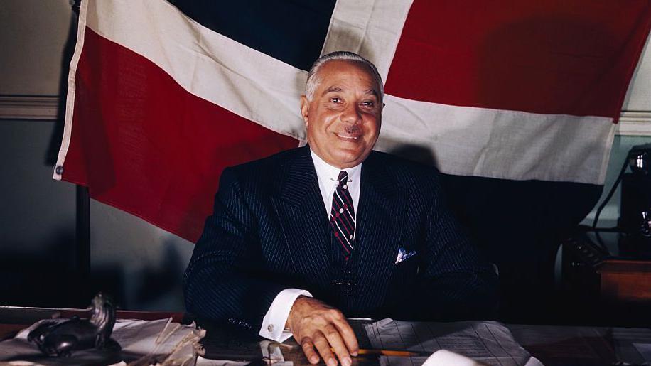 Rafael L. M. Trujillo, presidente de la República Dominicana, sonriendo con una bandera del país detrás.