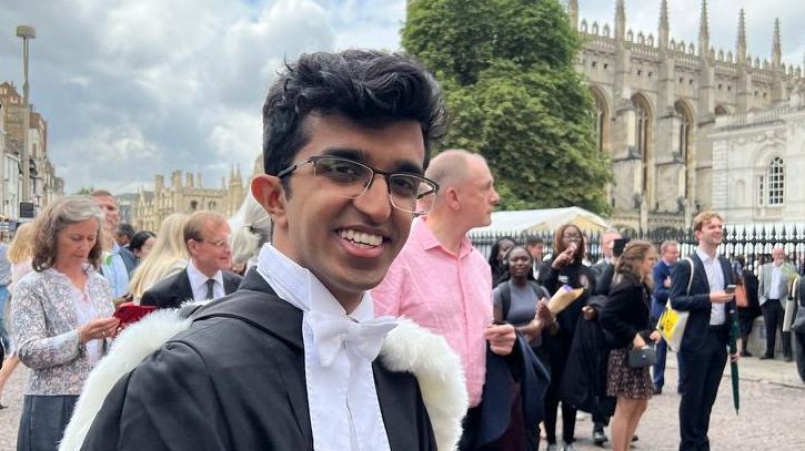 Ray vestindo uma beca de formatura em frente a uma das faculdades hist&oacute;ricas de Cambridge. Ele usa &oacute;culos e sorri para a c&acirc;mera