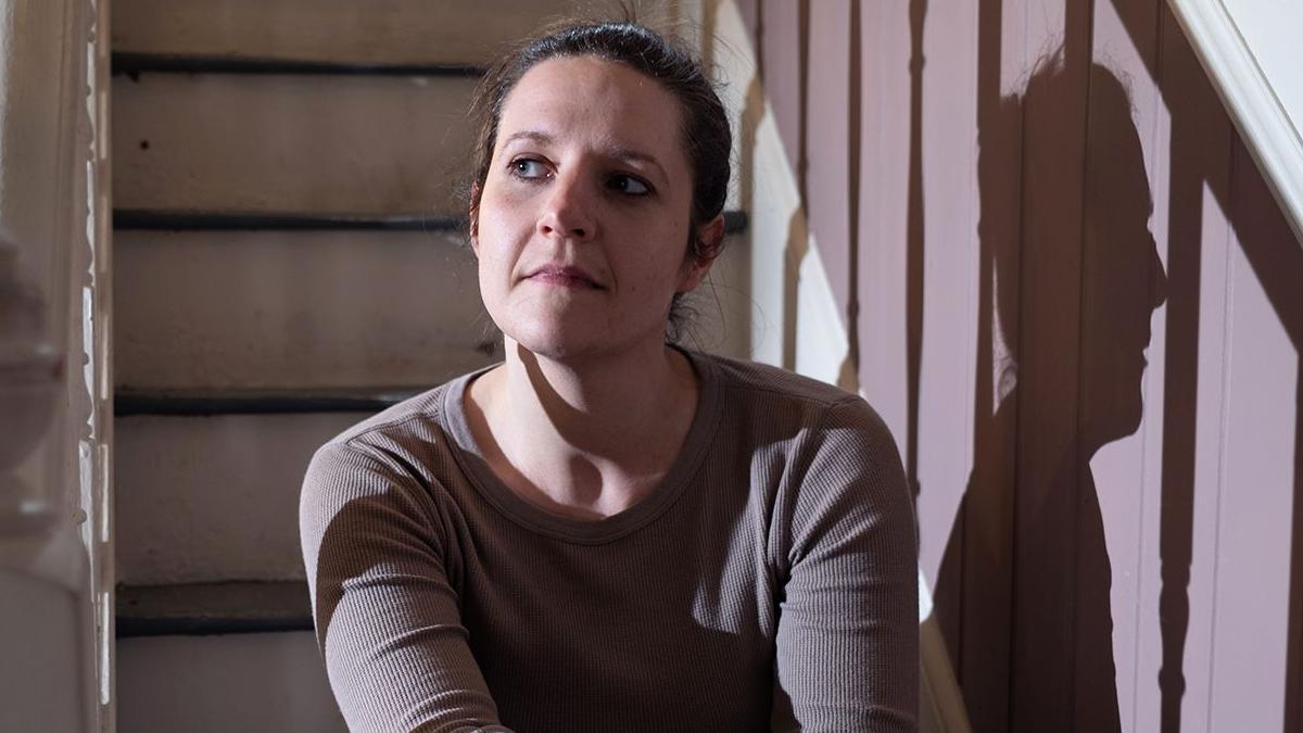 Ruth is sitting on an indoor staircase, illuminated by side lighting that casts a clear shadow on a paneled wall, looking slightly to the side with one arm resting on a raised knee and wearing a long-sleeved top and jeans.