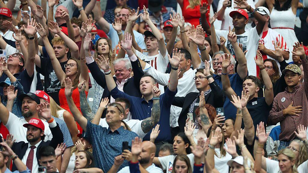 Um grupo de pessoas de diferentes idades e raças levanta os braços em louvor durante o evento em memória de Charlie Kirk.