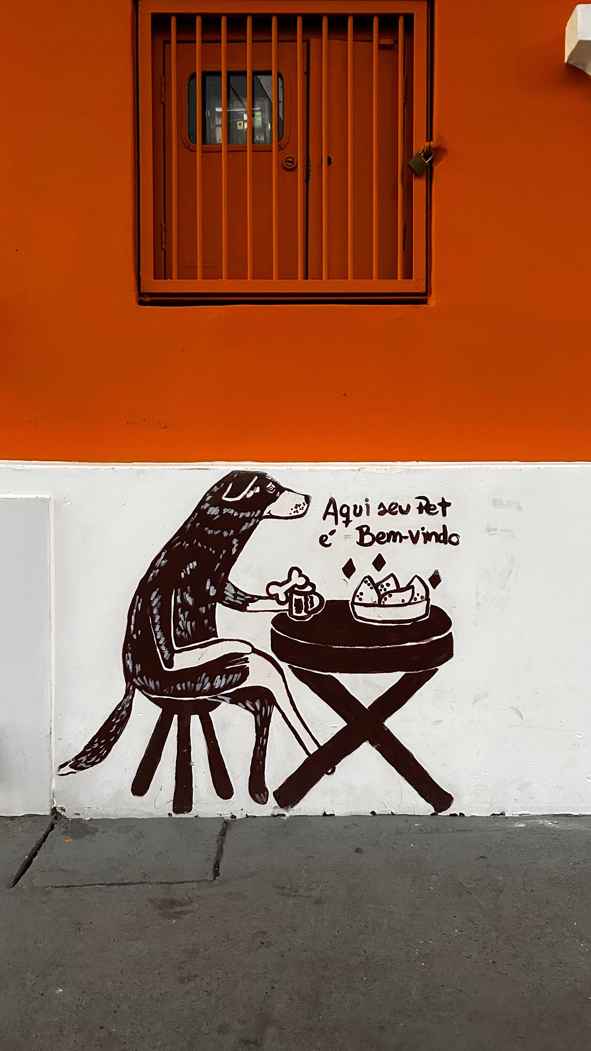 Pintura feita à mão na parede de bar em Porto Alegre, Rio Grande do Sul, mostra um cachorro de pernas cruzadas sentado em uma mesa de bar servida com um osso, uma bebida e uma porção de coxinhas. Acima se lê: