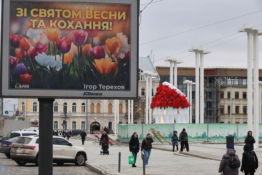На міській площі встановлено великий рекламний щит із кольоровими тюльпанами та написом: «ЗІ СВЯТОМ ВЕСНИ ТА КОХАННЯ!». Поруч розміщена велика декоративна інсталяція у вигляді букета червоних троянд. На площі ходять перехожі, видно припарковані автомобілі. У тлі видно будинки з вибитими вікнами, закритими фанерою.