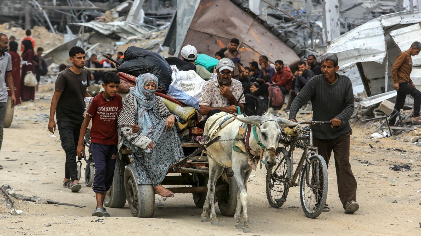 Un grupo de gente camina por un camino polvoriento con edificios arruinados a su alrededor. Con ellos, un burro tira del carro, en el que va sentada una mujer mayor y descalza. Junto al grupo camina un hombre que empuja una bicicleta.