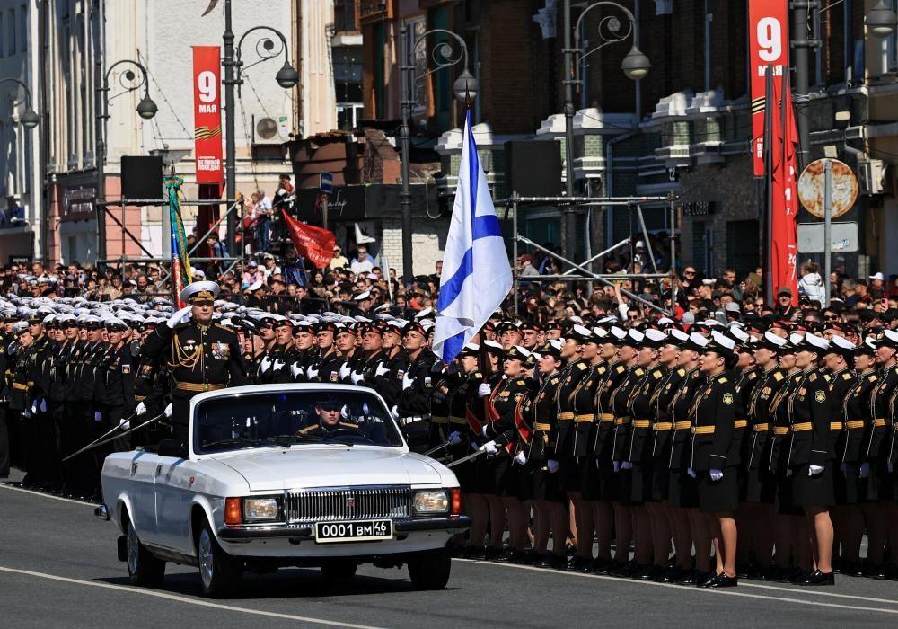 Un comandante militar ruso saluda mientras se encuentra de pie en un coche blanco descapotable conducido por otro soldado, en una carretera flanqueada por filas de militares. 