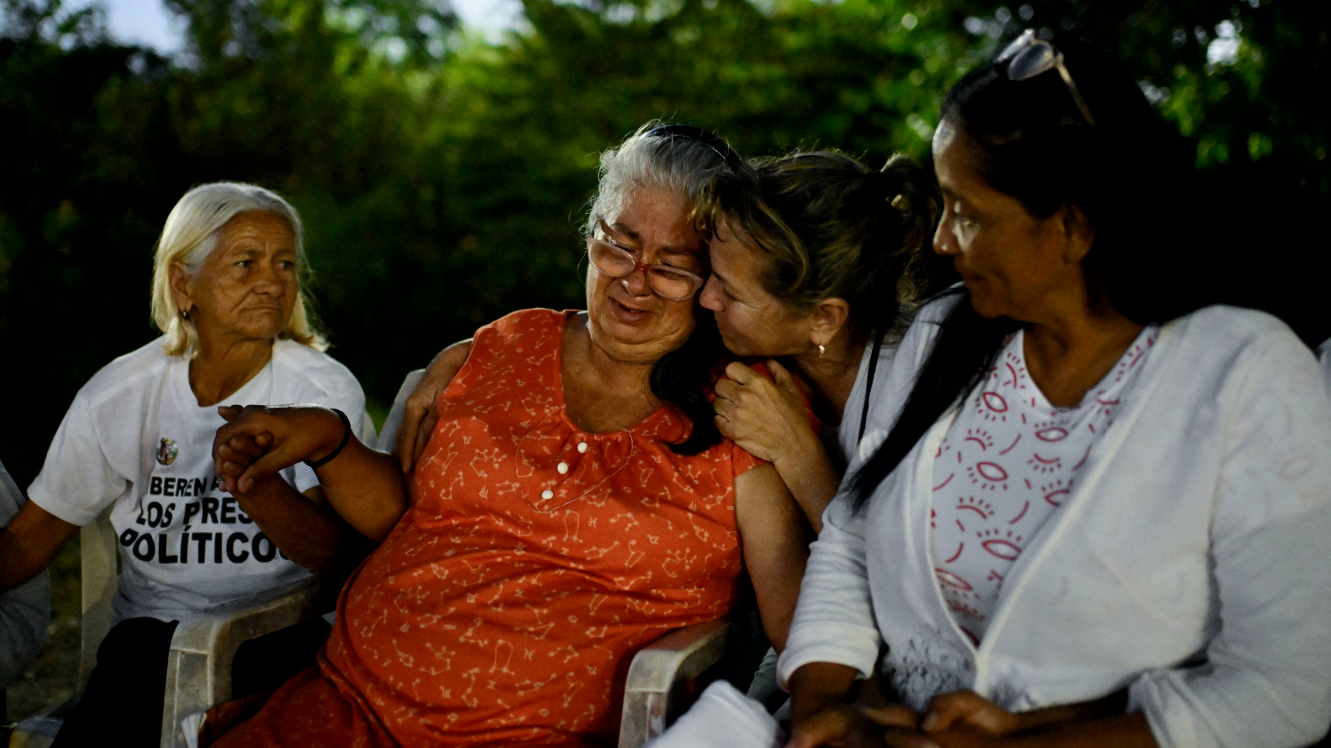 Un grupo de mujeres sentadas al aire libre; una de ellas llora y es consolada por otra que la abraza.