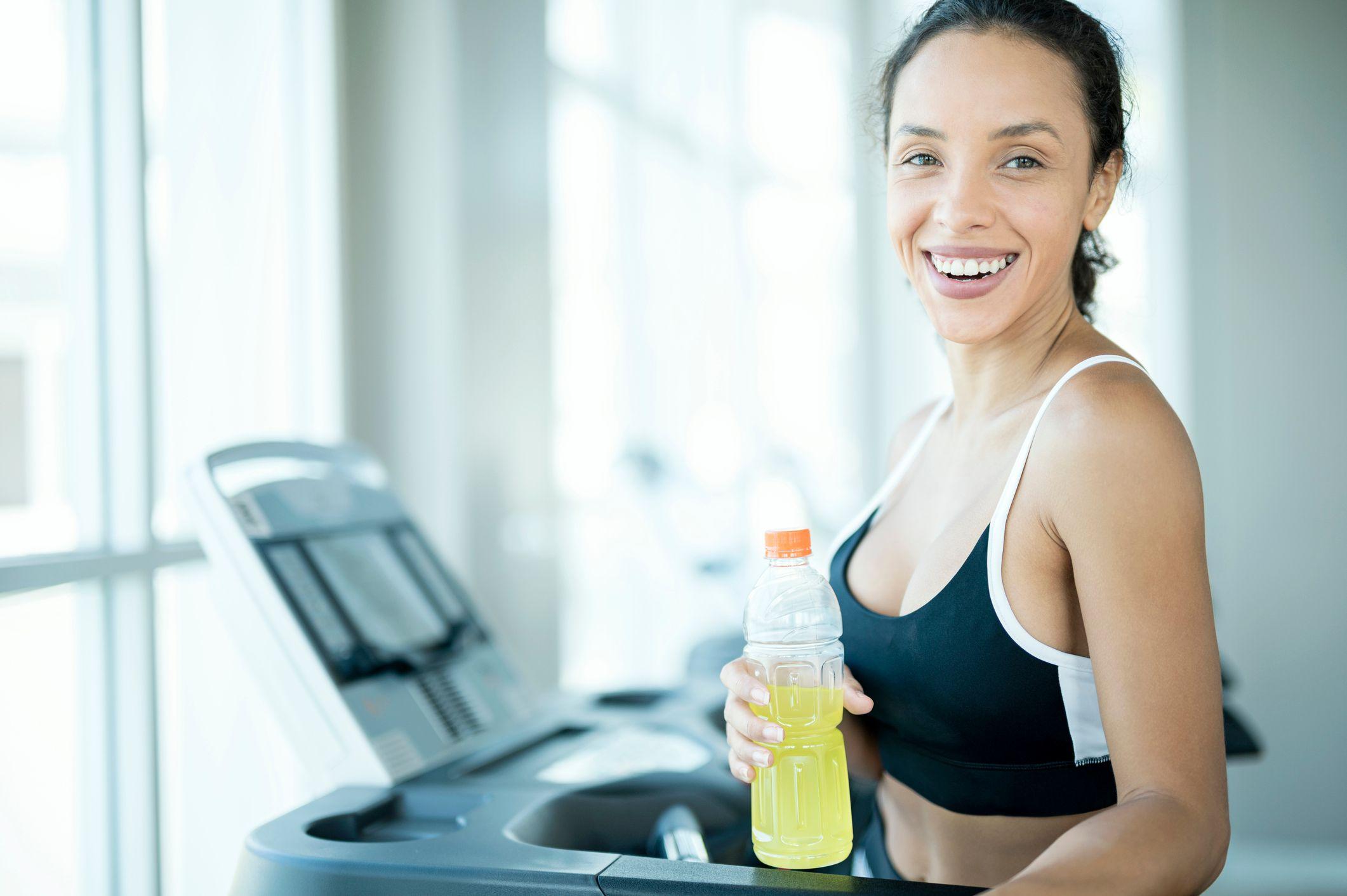Una mujer en ropa deportiva sonríe mientras sostiene una botella de plástico con una bebida energética de color amarillo fluorescente. 