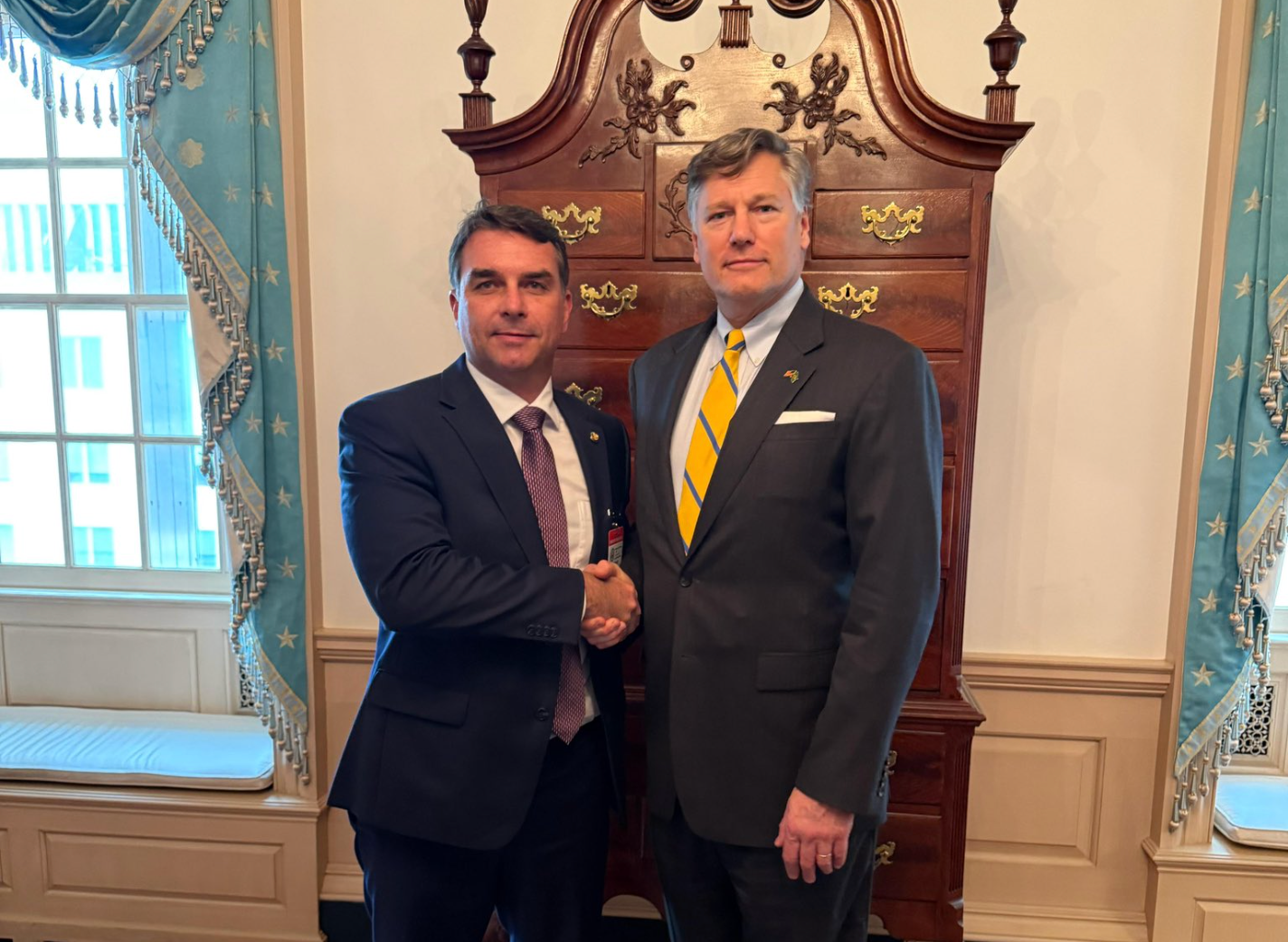 Foto com Fl&aacute;vio Bolsonaro (&agrave; esquerda) e Christopher Landau, &agrave; direita. Os dois apertam as m&atilde;os diante de um arm&aacute;rio de madeira em estilo cl&aacute;ssico. 
