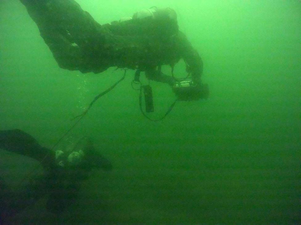 Dos miembros del equipo de buzos que buscan minas. Están  bajo el agua que tiene un color verde brillante.