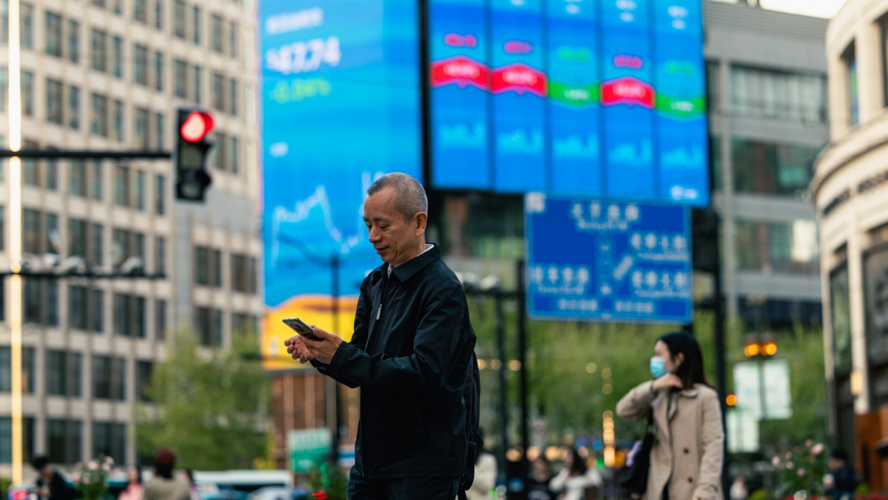 一名男子走在上海一條高樓林立的街道上，手裡拿著手機，其中一棟建築的正面顯示著當地交易所的業績