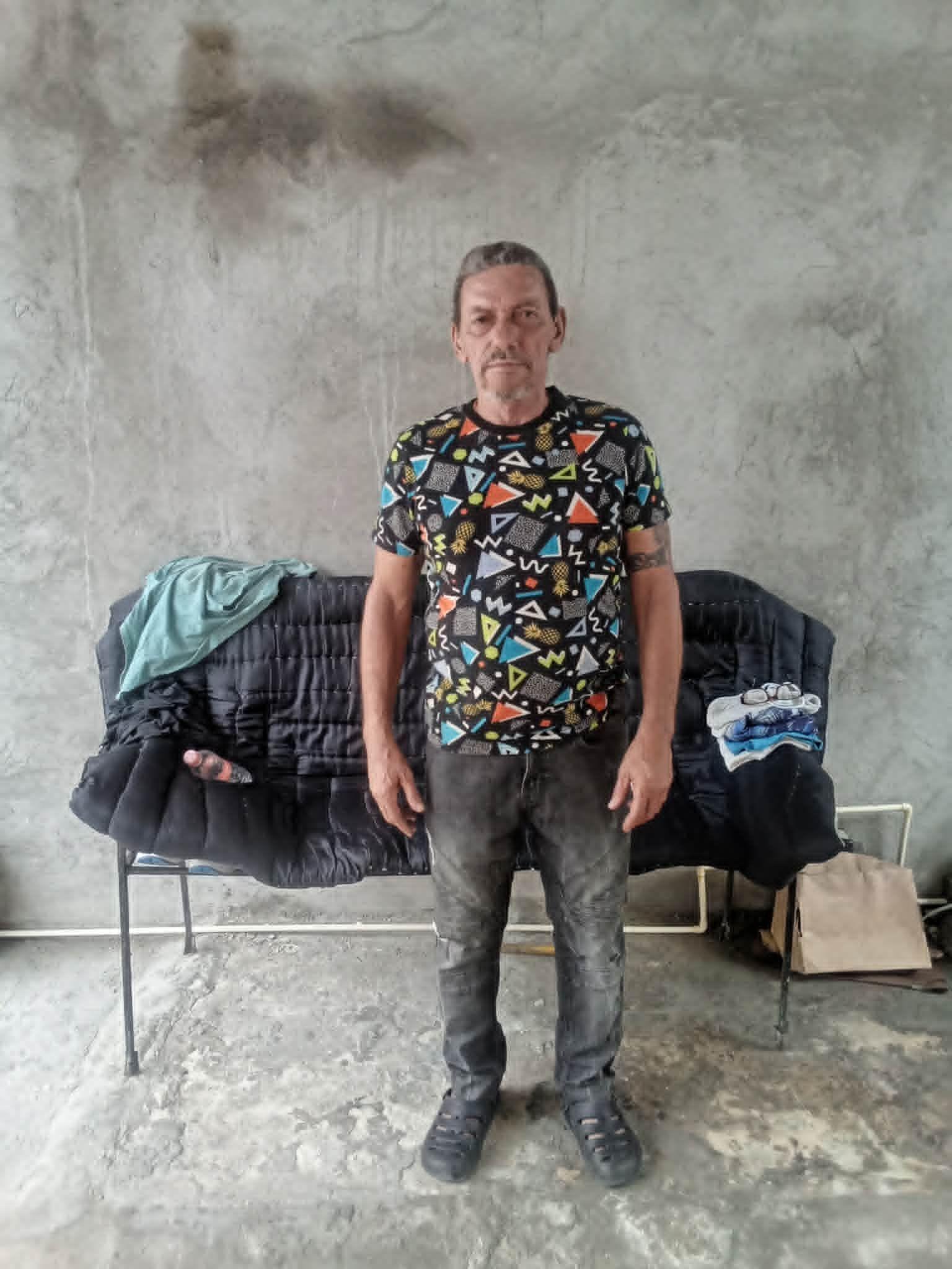 Lázaro Romero León mira a cámara de pie frente a una silla alargada, con jeans gris oscuros y una camiseta estampada, en un patio de cemento en Tapachula, Chiapas, México.
