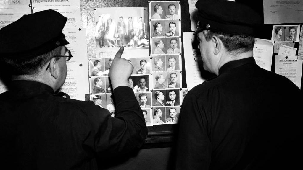 Dois policiais do Brooklyn apontam para a fila de membros da gangue Murder Inc., por volta de 1940, na delegacia de polícia do Brooklyn, em Nova York.