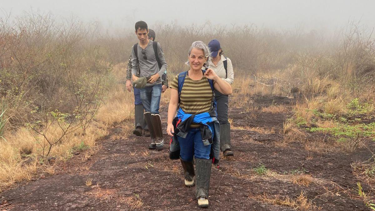 Ana sorrindo ao caminhar em área com arbustos; há outras pessoas caminhando atrás dela
