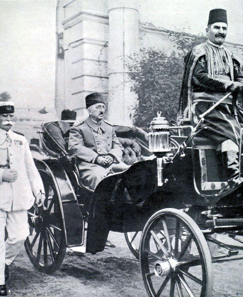 Mehmed VI, lleva un abrigo, está sentado en carruaje que transita por la calle. A su lado, camina un funcionario vestido de blanco