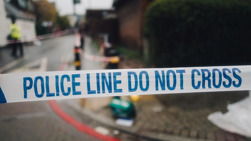 Garis polisi berwarna putih dan biru memisahkan tempat kejadian perkara.