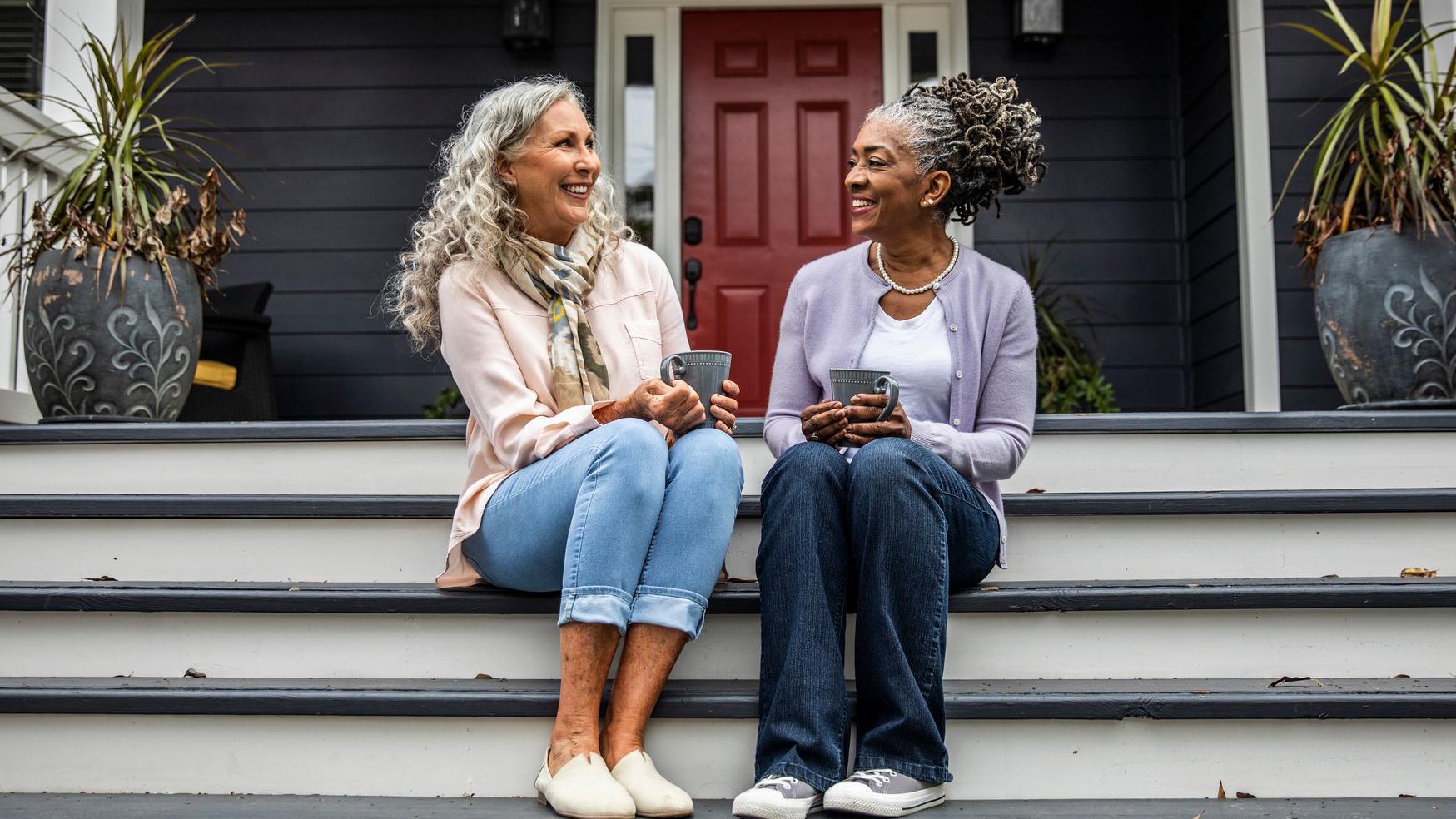 Dve starije žene sede na tremu držeći šoljice kafe u rukama i razgovarajući