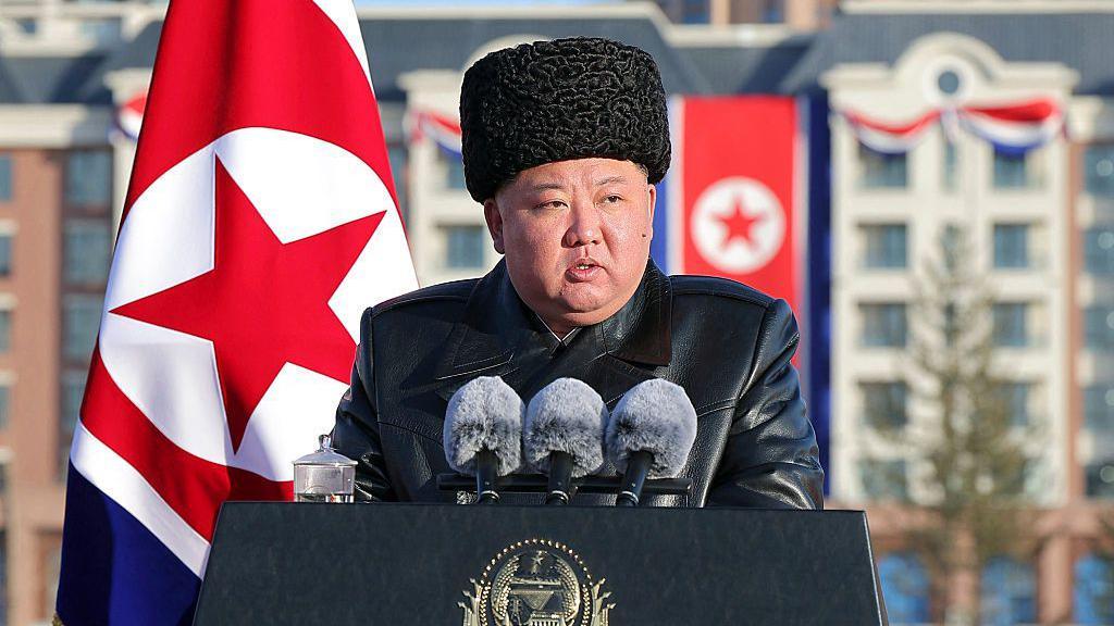 Kim Jong-un habla desde un atril con tres micros. Lleva un gorro y tiene la bandera de su país detrás. 