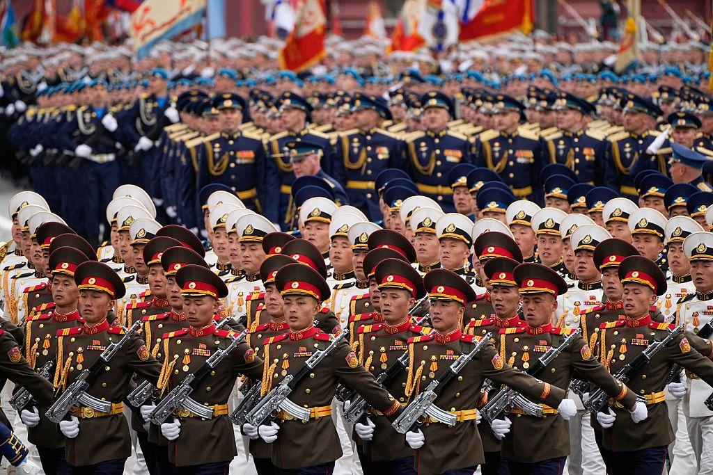 Soldados norcoreanos desfilan en filas portando armas. Los soldados de las primeras filas visten uniformes de color caqui con galones dorados y rojos. Más atrás se encuentran otros soldados norcoreanos con uniformes blancos con galones dorados y negros. Detrás de ellos hay filas de soldados rusos.
