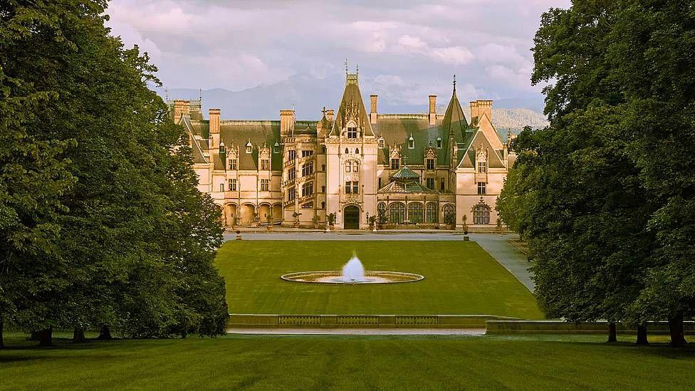 Vista panorámica de la mansión Biltmore que se ve tras un gran jardín con árboles y una fuente en el centro  