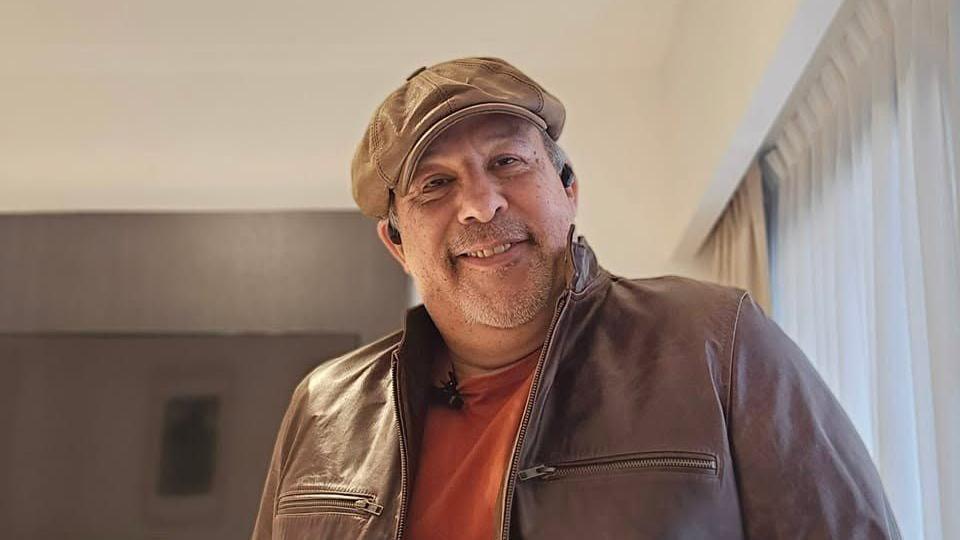 Roberto Guido, um homem de meia idade, vestindo um bon&eacute; de couro e jaqueta, ambos na cor marrom, com uma camiseta laranja por baixo. Ele sorri para a c&acirc;mera e tem uma barba rala grisalha