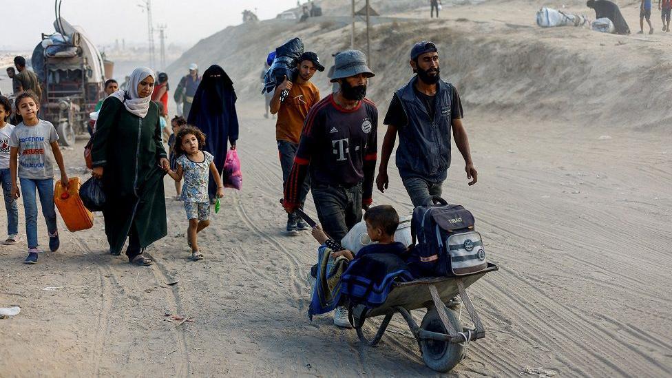 Hombres, mujeres y niños caminan con sus pertenencias sobre una zona arenosa. Un hombre empuja una pequeña carretilla donde está un niño con un bolso y pertenencias 