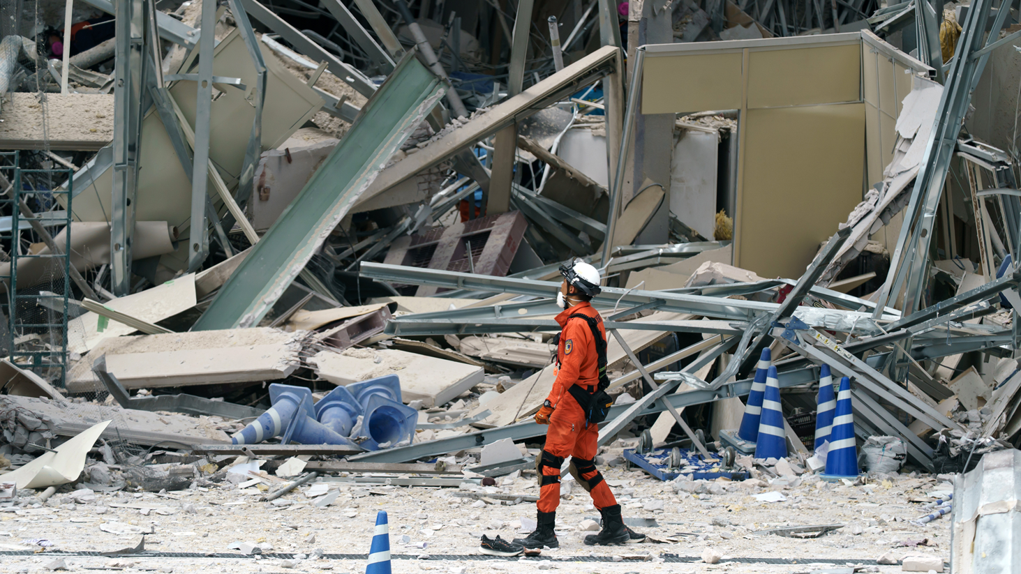 Un bombero, con uniforme anaranjado, botas negras, casco y mascarilla blancos, pasa por el lado de los escombros de una infraestructura

