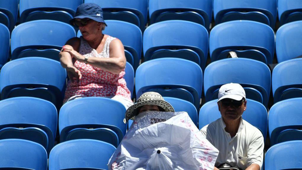 Una espectadora usa una sombrilla para protegerse del sol mientras otra se aplica protector solar durante el partido de tercera ronda de individuales masculino entre Gilles Muller de Luxemburgo y Pablo Carreño Busta de España en el quinto día del torneo de tenis Abierto de Australia en Melbourne el 19 de enero de 2018.