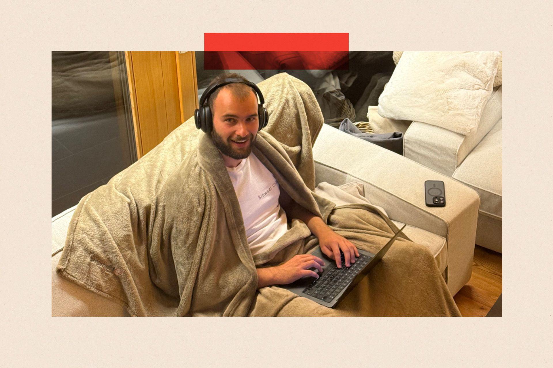 Magnus Müller envuelto en una manta, sentado en un sofá y trabajando con su ordenador portátil.