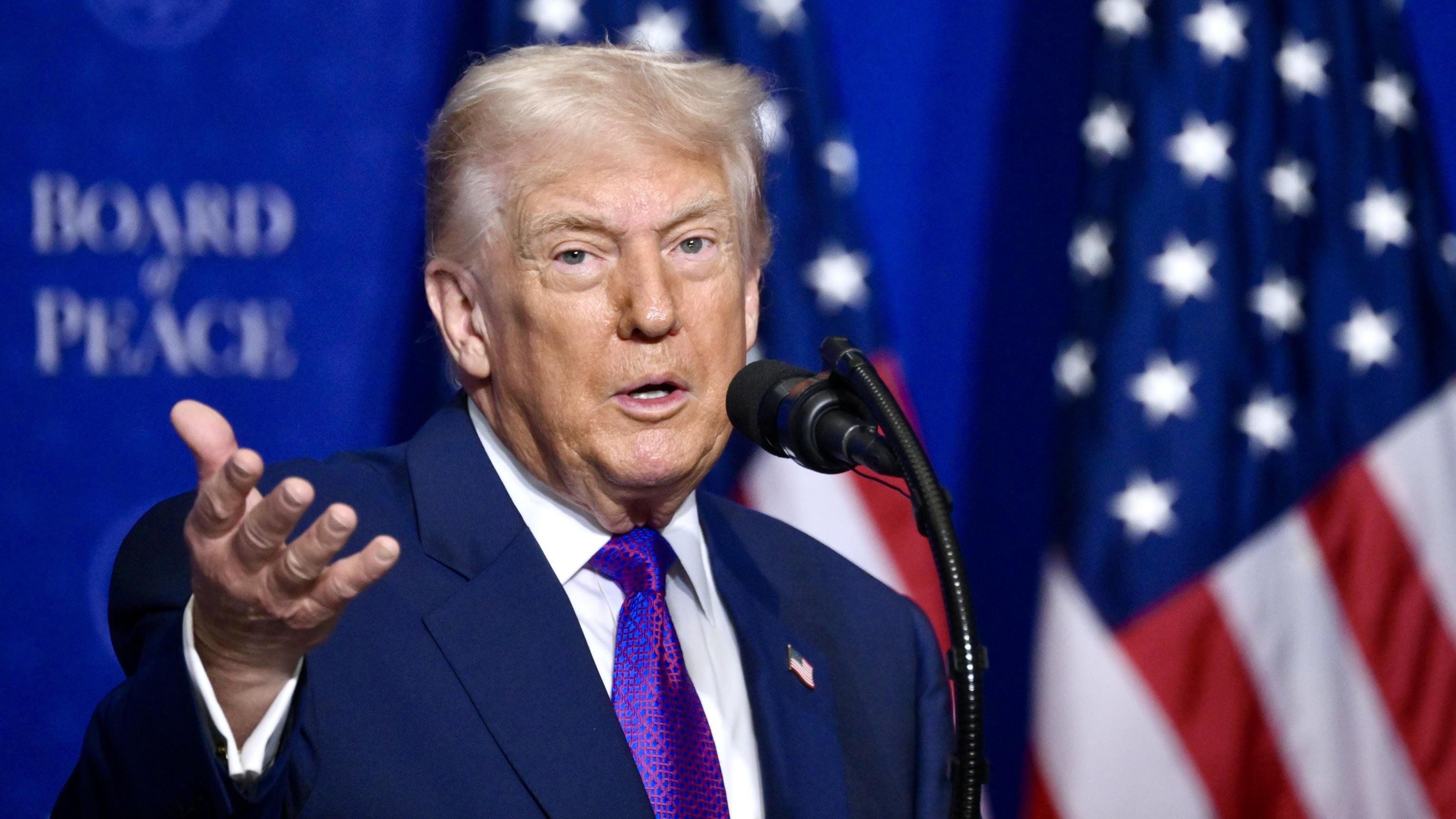 ترامب يتحدث خلال اجتماع مجلس السلام في واشنطن العاصمة في 19 فبراير/شباط 2026. وهو يرفع يده اليمنى أثناء حديثه عبر الميكروفون، والعلم الأمريكي خلفه.