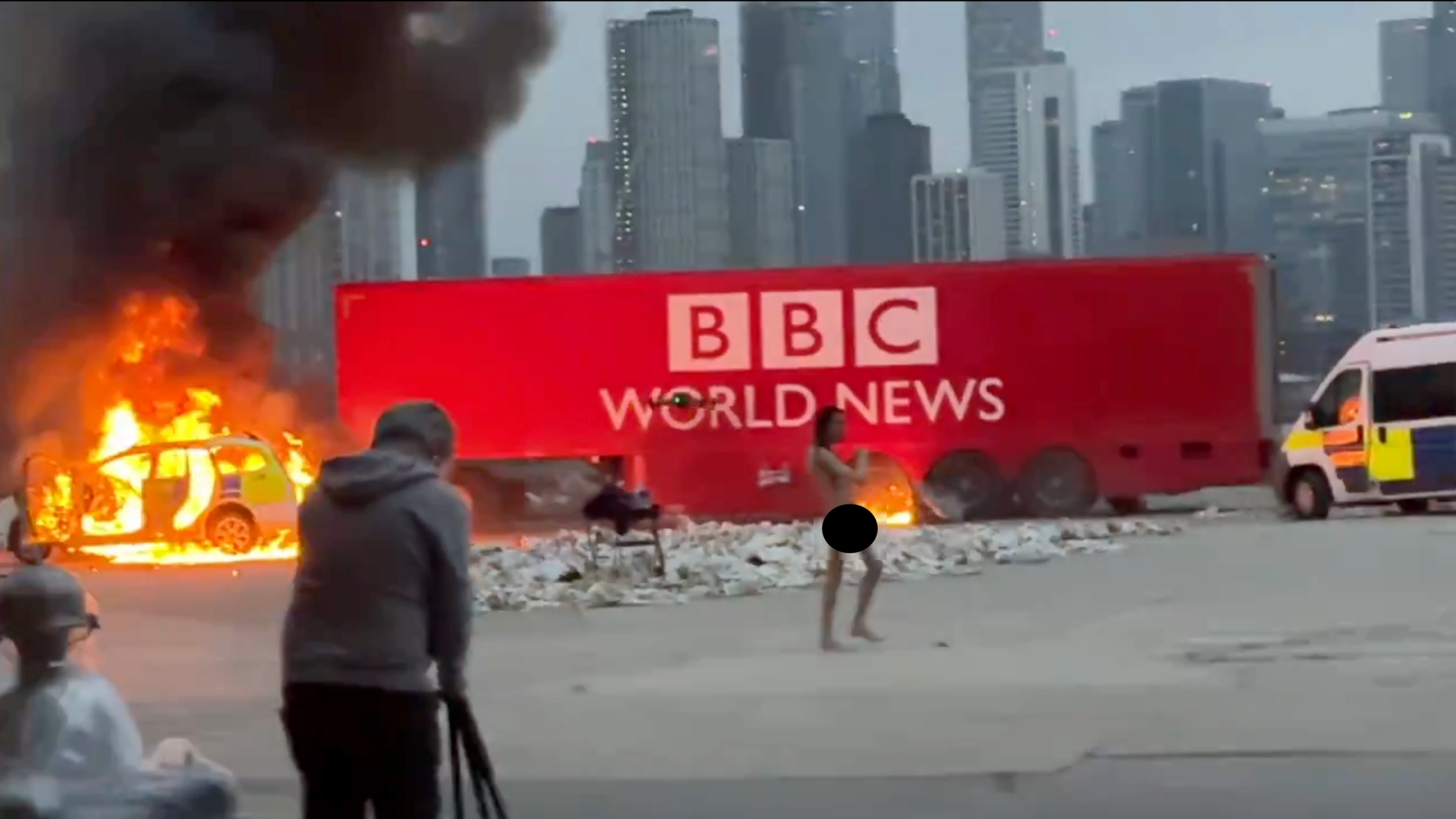 Uma cena de um vídeo que mostra Jhaj, nu, mas com sensores, em pé em frente a um grande caminhão vermelho com o logotipo da BBC World News, e parte do caminhão em chamas. Um cinegrafista pode ser visto em primeiro plano.