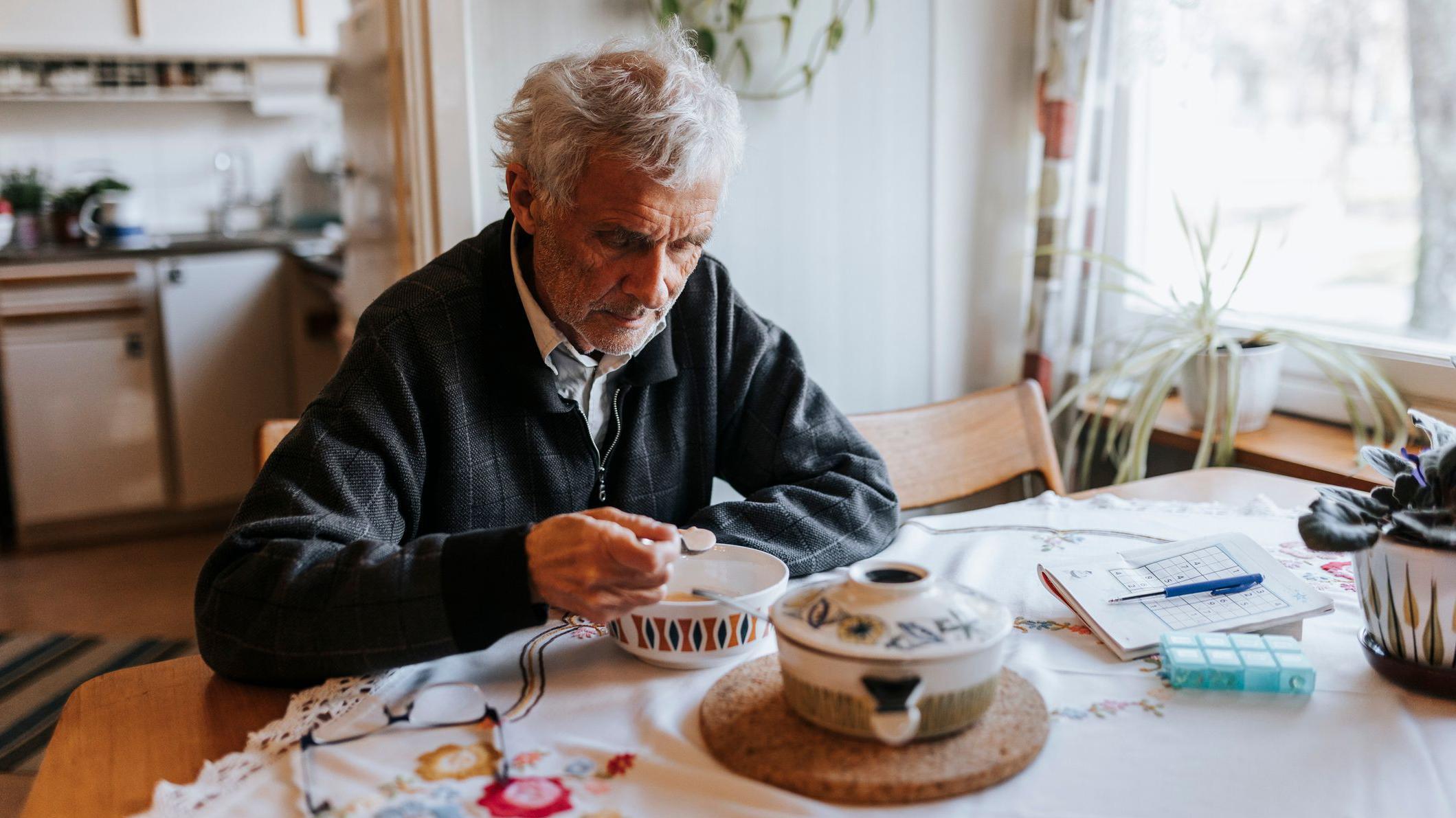 회색 머리의 노인이 집 안 식탁에 앉아 그릇에 담긴 시리얼을 먹고 있다. 