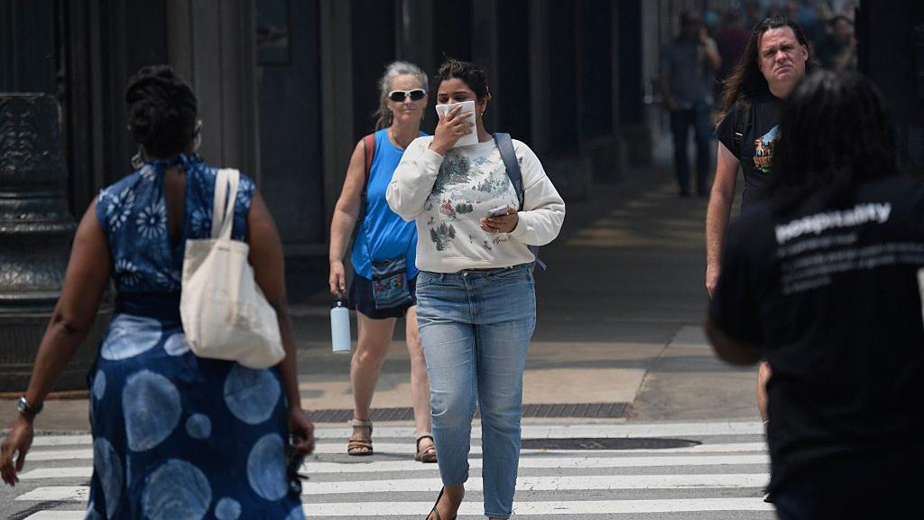 Una mujer vestida con una sudadera blanca y jeans se cubre el rostro con un pañuelo mientras cruza una calle de la ciudad, junto con otros peatones. 