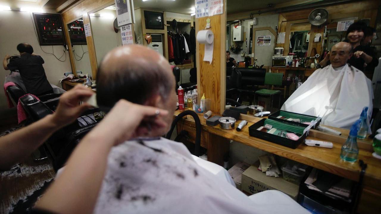 一位正在理髮店理髮的禿頂老人的背影，透過他面前的鏡子可以看到他的臉 。