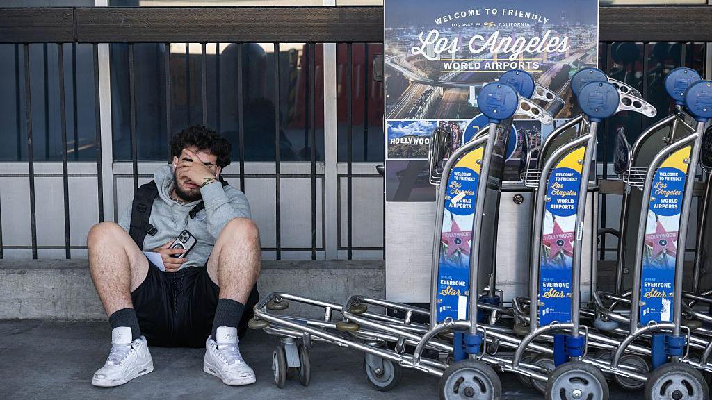 Un hombre sentado en el piso junto a varios carritos para transportar maletas típicas de los aeropuertos. El hombre tiene la mano sobre el rostro con un gesto que denota agotamiento.